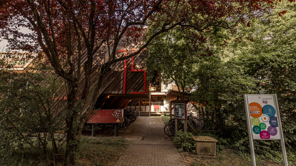 Foto des Eingangs vom Bauhäusle. Ein gepflasterter Weg für durch eine Reihe Bäume und unter einem verwinkelten Holzbau zur Eingangs-Terrasse des Bauhäusle.