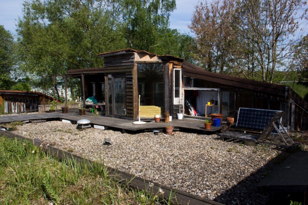Foto auf der Dachterrasse. Eine Kies-Fläche im Vordergrund und eine Holzfassade mit kleinem Wintergart daran anschließend. Im Hintergrund Bäume