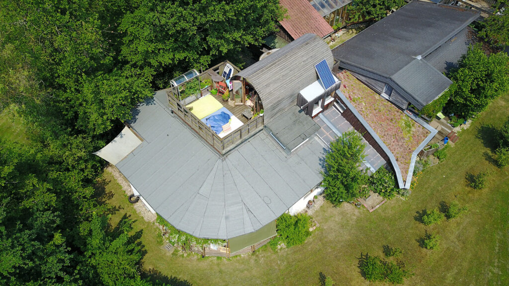 Drohnen-Aufnahme des hinteren Teils des Gebäudes, mit dem aufgesetzten Holz-Tonnen-Zimmer. Darum herum Bäume und grüne Wiese
