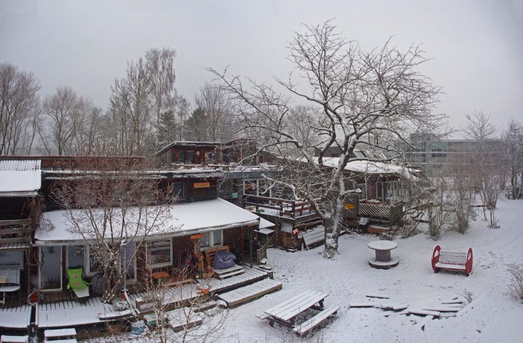 Luftaufnahme des Südgartens im Winter. Ein verwinkelter Holzbau mit schneebedeckter Wiese und Dach