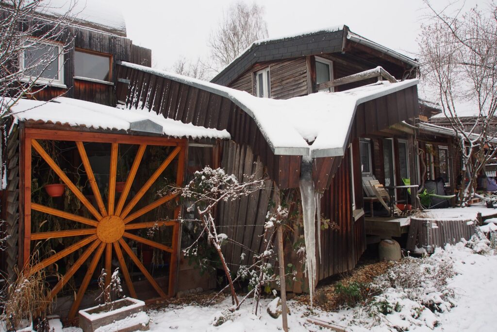 Ein verwinkelter Holzbau mit schneebedecktem Dach und Wiese. Ein großer Eiszapfen hängt von einer Dachecke