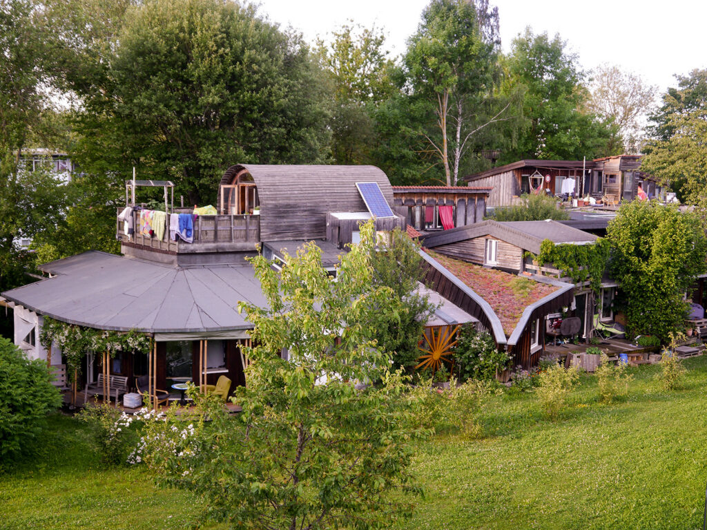 Luftaufnahme des Hauses von Südseite. Eine verwinkelte Holzbau-Konstruktion mit dem aufgesetzten Holz-Tonnen-Zimmer. Im Vordergrund eine grüne Wiese und im Hintergrund hohe Bäume