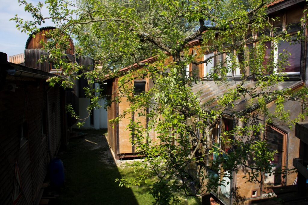 Aufnahme des Innenhofs. Ein Holzbau mit der gläsernen Fassade des Wintergartens und eine Baum im mittigen Vordergrund