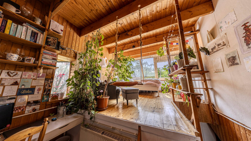 Weitwinkel-Foto aus einem Zimmer. Man sieht rechts eine Schreibtischecke mit Wandregal und dekorierter Wand. Mittig ist eine erhöhte Ebene mit Sessel, Schaukel und Bett die zu einem großen Schrägdach-Fenster führt.