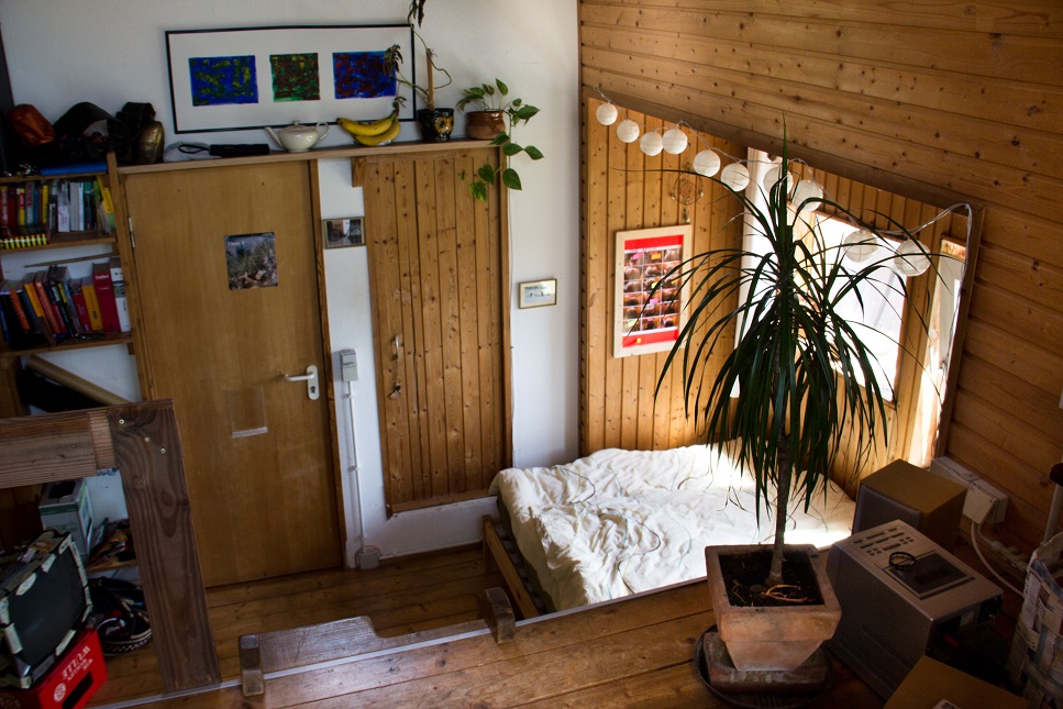 Foto aus einem Zimmer. Von der zweiten Ebene schauen wir auf die erste, wo rechts ein in eine Nische in der Holzwand ein Fenster und Bett eingelassen sind. Links ist die Zimmertür an einer dekorierten Wand mit Regal