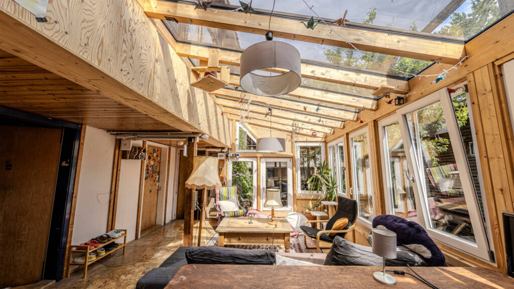 Foto im Wintergarten. Ein fast vollständig verglaste Holzkonstruktion mit einem Überbau und Zimmertüren an einer Wand links. Im Wintergarten stehen mehrere Sessel ein Tisch und einen alter Stehlampe.