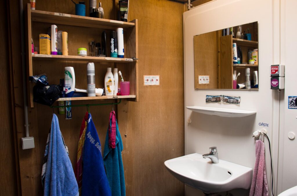 Fot vom Bad. Rechts ein Waschbecken mit Spiegel und links an der Holzwand ein Holzregal mit Bad-Utensilien und hängenden Handtüchern.