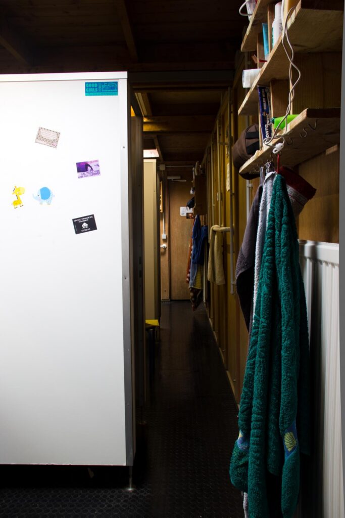 Foto vom Bad. Entlang der Toiletten-Kabinen und Duschen auf der linken Seite. Rechts an der Holzwand Holzregale mit Bad-Utensilien