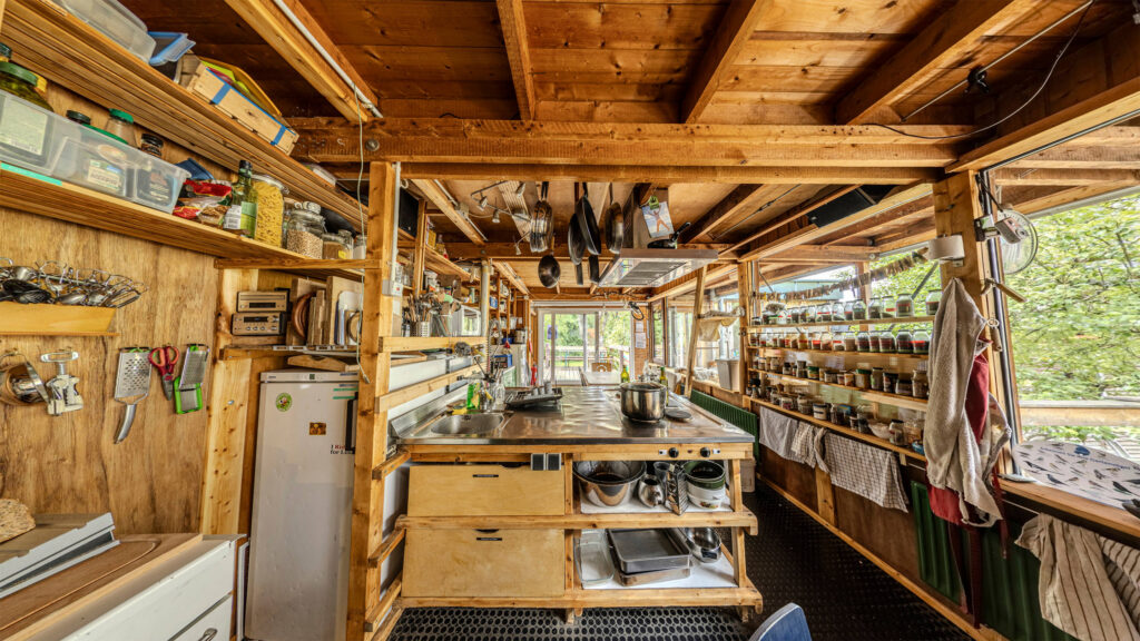 Foto der hinteren Küche. Eine Holkonstruktion mit einer vollen Regalfassade links, einer Kücheninsel mit Waschbecken und Herd in der Mitte und eine Glasfassade mit Gewürzregal rechts. Im Hintergrund eine große Terrassentür.