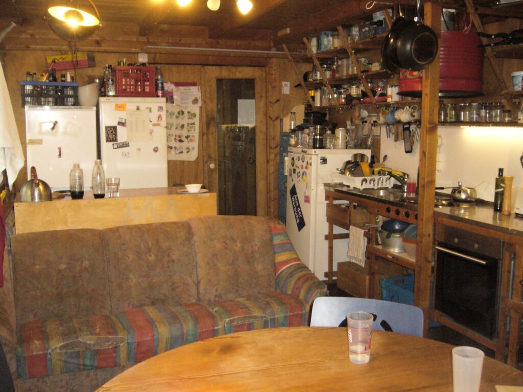 Foto der vorderen Küche. links im Hintergrund Külschränke und ein Tresen, rechts eine Küchenzeile mit vielen, vollgestellten Wandregalen darüber und Im Vordergrund eine bunte Couch und ein Küchentisch