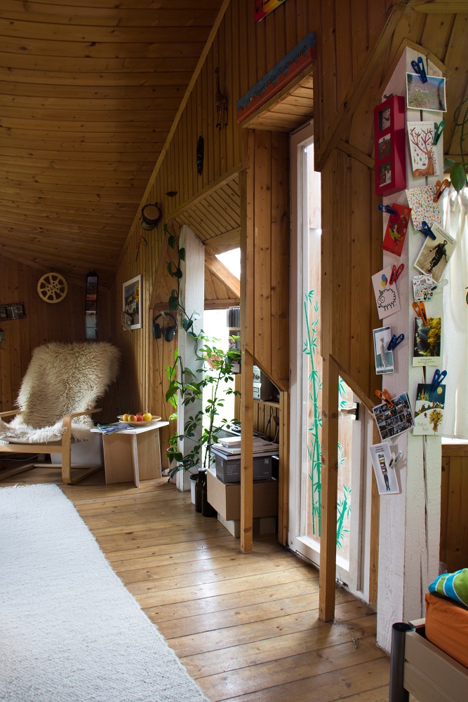 Foto aus einem Zimmer. Ein winkeliger Raum mit Holzwand und großen Fenstern. An der Wand Deko und Pflanzen und einen flauschiger Sitzsessel im Hintergrund.