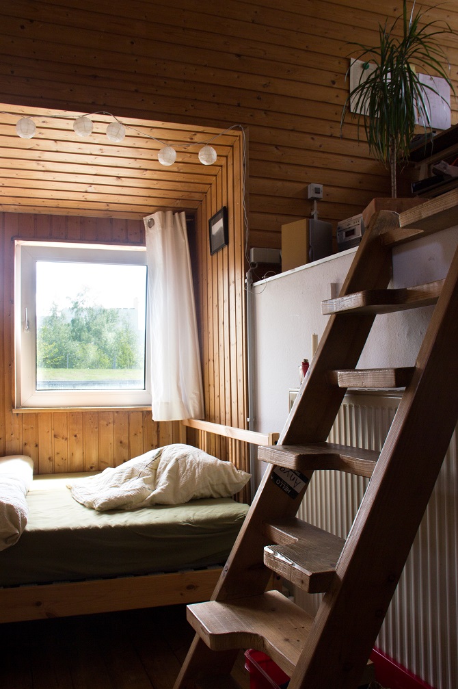 Foto aus einem Zimmer. In die Holzwand eingelassene Nut mit Fenster und Bett. Rechts eine Holz-Samba-Treppe die auf eine zweite Ebene führt.