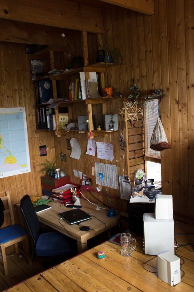 Foto aus einem Zimmer. Eine Holzzimmer, in dem auf einer niedrigen Ebene eine belebte Schreibtisch-Ecke mit Wandregal ist.