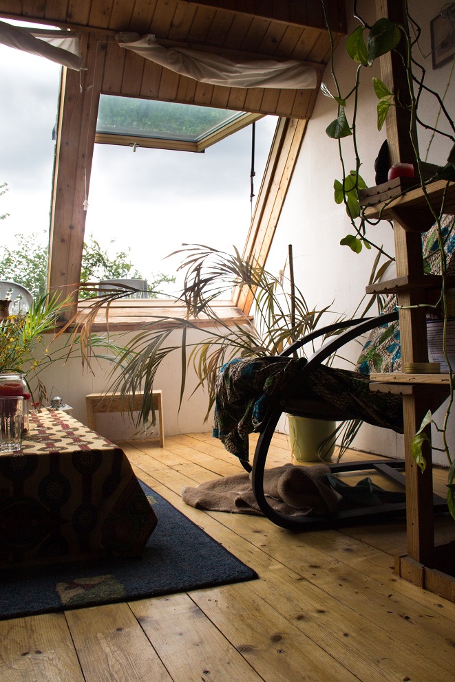 Foto aus einem Zimmer. Unter einem großen Schrägdachfenster stehen mehrere Pflanzen, einen gemütlicher Stuhl und ein kleiner niedriger Tisch mit bunter Tischdecke