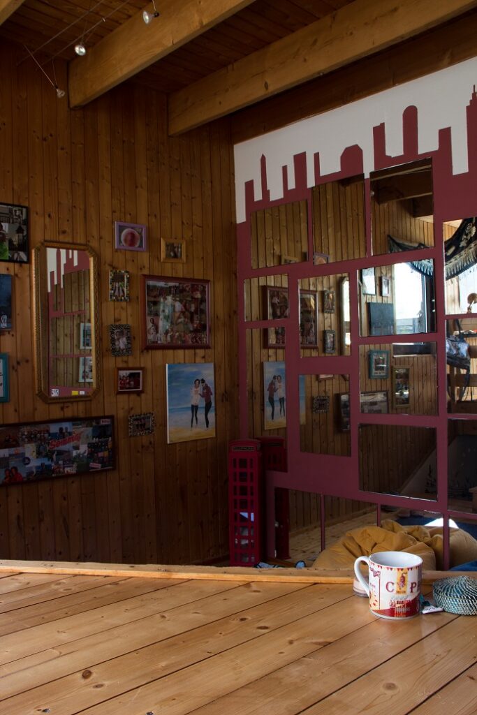 Foto aus einem Zimmer. Eine Holzwand-Ecke mit Fotos und Gemälden an der linken Wand und mehreren Spiegelflächen an der rechtenWand