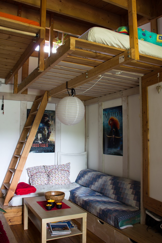 Foto aus einem Zimmer. Unten ist eine gemütliche Sitzecke, Oben ist unter der Decke ein Lattenrost mit Matratze als zweite Ebene zu der links eine Leiter führt.