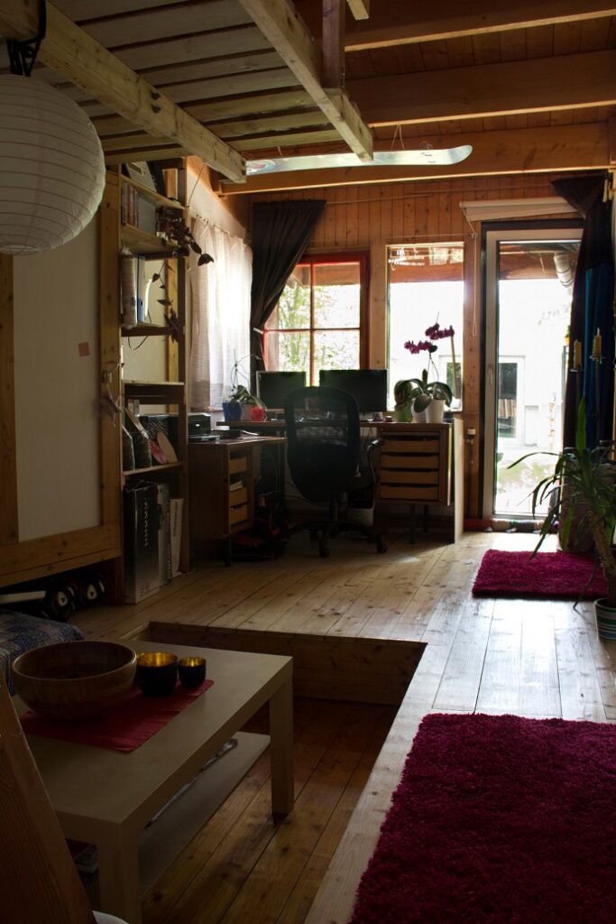 Foto aus einem Zimmer. Ein langgezogenes Holzzimmer mit einer kleinen in den Boden eingelassenen Sitzzecke mit Tisch. Im Hintergrund steht ein Schreibtisch vor der Fensterfassade