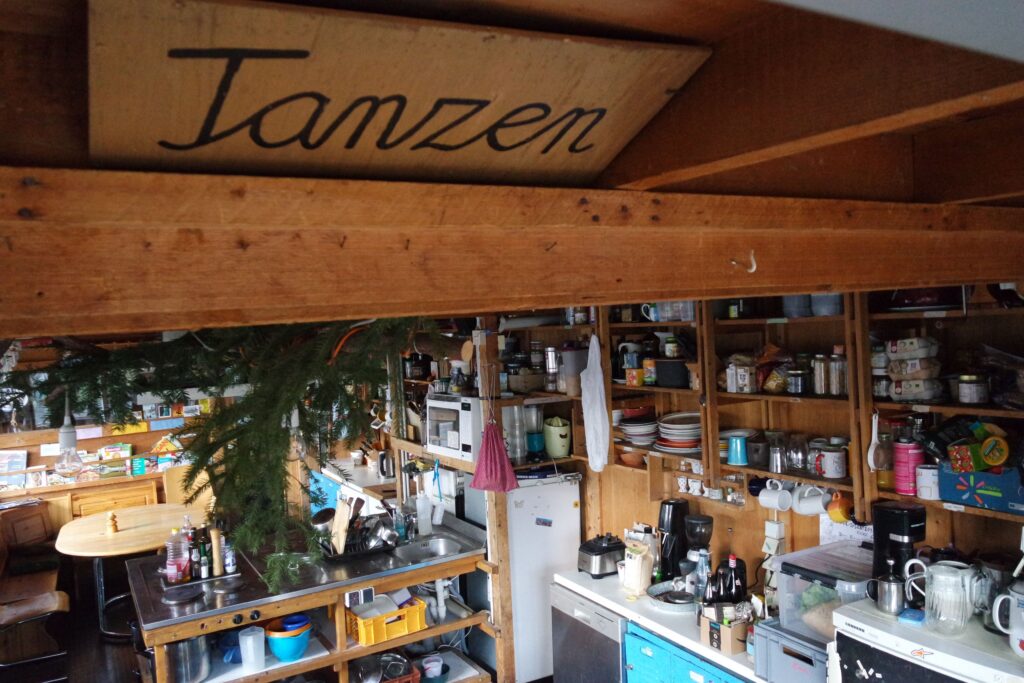 Foto von der bunt eingerichteten hinteren Küche mit der Kochinsel in der Mitte. Unter der Decke klemmt eine Holzschild mit dem Wort "Tanzen" darauf