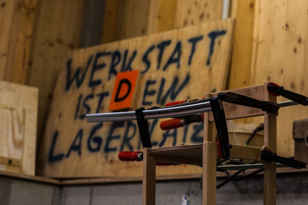 Nahaufnahme aus der Werkstatt, wo ein Stuhl mit Schraubzwingen trocknet. Im Hintergrund ein großes Handgeschriebenes Schild "Werkstatt ist dein Lagerplatz" wobei das D nachträglich aufmontiert wurde