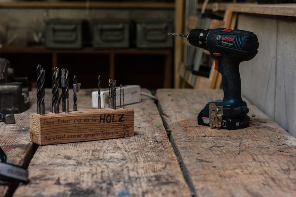 Nahaufnahme von der Werkbank in der Werkstatt, auf der eine Akku-Schrauber mit mehreren Holzbohrer-Aufsätzen daneben steht