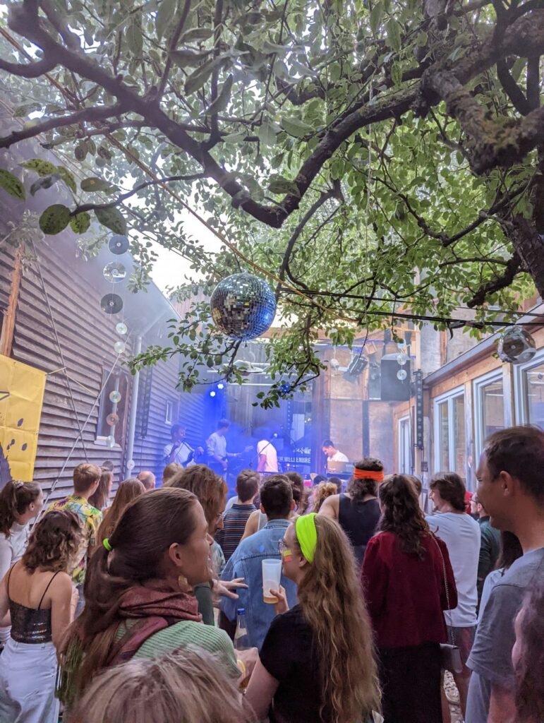 Aufnahme aus dem Innenhof bei einer Party. Mit Holzwänden links und rechts steht in der Mitte eine frohe Menge Menschen die der Band auf der Bühne im Hintergrund bei ihrem Auftritt zusieht und zuhört. Am oberen Bildrand ragt ein großer Baum ins Bild, in dem mehrere Diskokugeln hängen.
