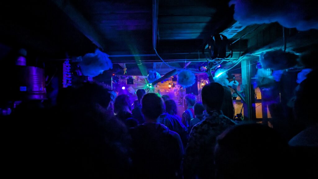 Dunkle Aufnahme von einer Menge Menschen die in der vorderen Küche einer Band beim Spielen zuschaut. Die Beleuchtung ist Blau mit Laserstrahlen und kommt nur von der Bühne aus.