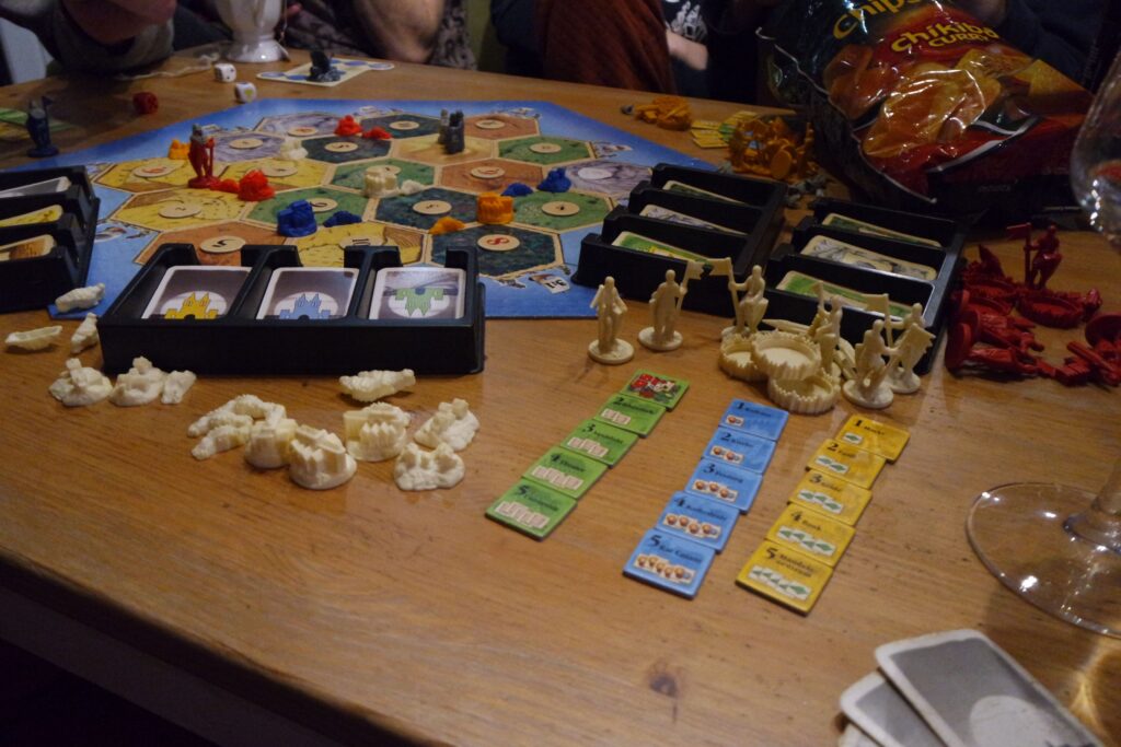 Nahaufnahme von einem Holztisch auf dem Siedler von Catan gespielt wird. Buntes Spielbrett mit Figuren und Karten, eine offene Tüte Chips rechts oben