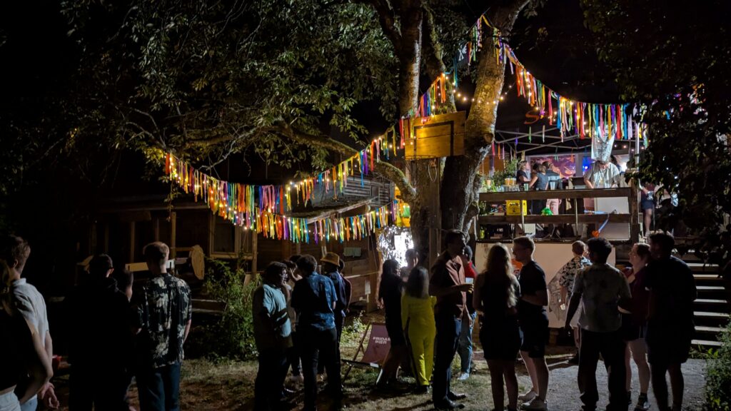 Nachtaufname des Südgartens bei einer Party. Viele Menschen im Gespräch, zwischen den Bäumen hängen bunte Girlanden, rechts im Hintergrund die Terrasse mit Bierbar