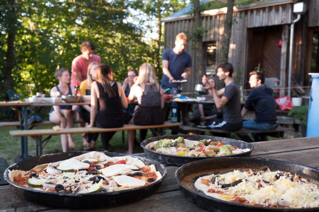 Im Vordergrund belegte runde Pizzen, die auf den Ofen warten, im Hintergrund unscharf eine Gruppe Studierende die im Garten gemeinsam an einem langen Tisch sitzen und Essen