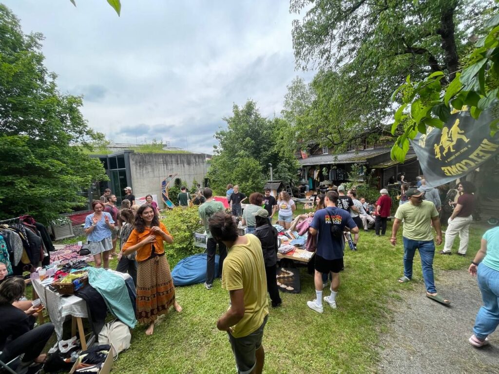 Foto von einem gut besuchten Flohmarkt im Südgarten des Hauses. Auf der grünen Wiese sind mehrere Standflächen mit Kleidung und Gegenständen aufgebaut und viele Menschen stehen dazwischen